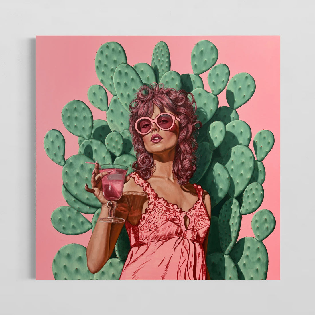 A canvas print featuring a stylized illustration of a woman with cacti in the background, drinking from a glass, set against a pink and green color scheme.