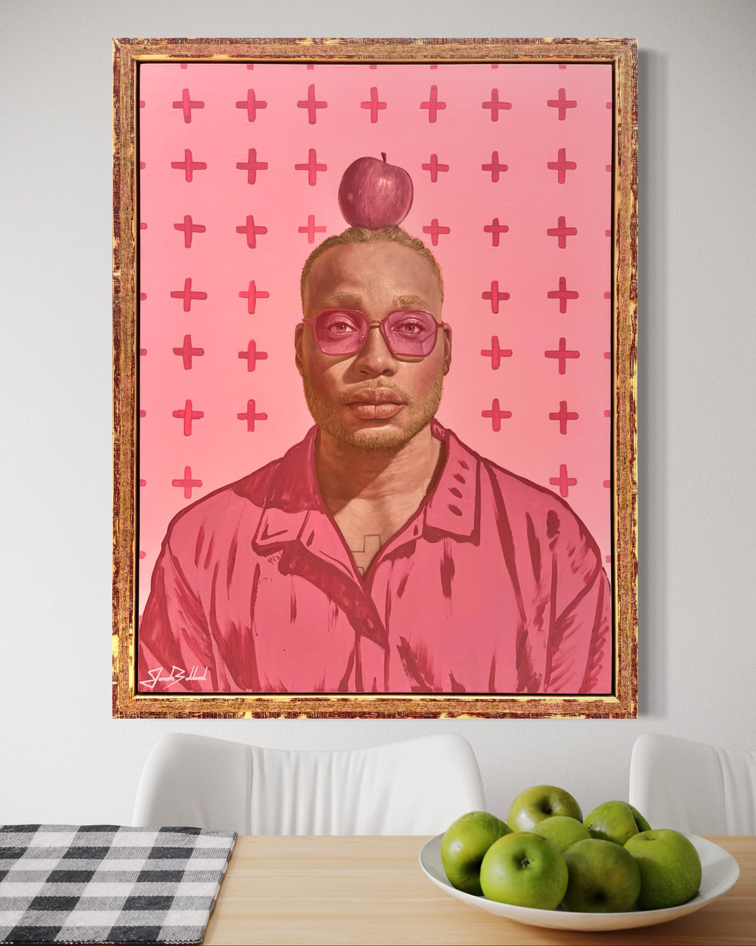 Framed artwork of a man with an apple on their head against a pink cross-patterned background, displayed in a room with a table and apples.
